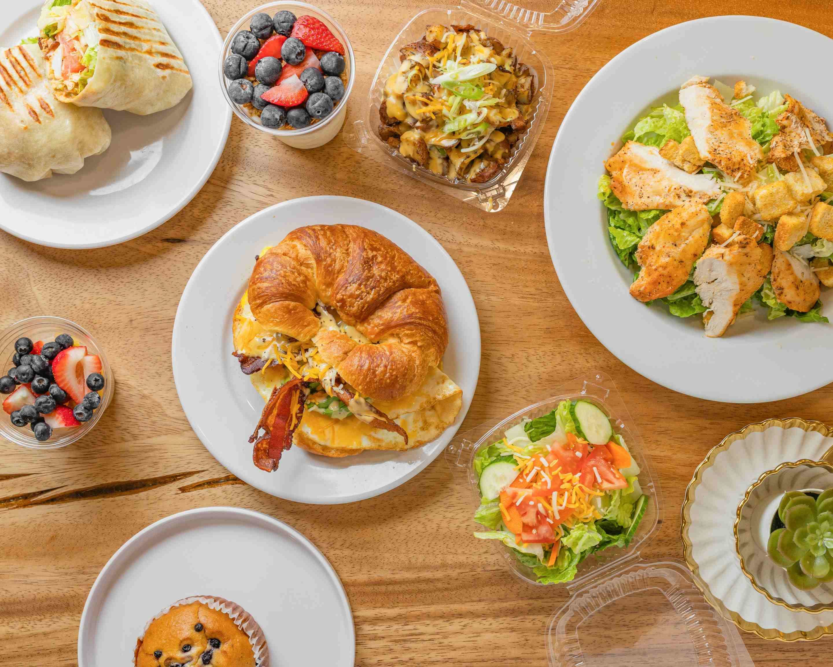Overhead spread of Daily Dose breakfast and lunch favorites including a bacon croissant sandwich, grilled chicken caesar salad, wraps, garden salad, and fresh fruit cups